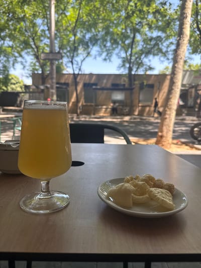A close-up, outdoor shot features a glass of pale yellow liquid, likely beer, and a small white plate filled with golden-brown puffed snacks, possibly pork rinds, resting on a wooden table. In the background, the blurred scene depicts a street lined with trees and buildings, suggesting an urban or suburban setting. The lighting indicates daytime, with sunlight filtering through the leaves. The presence of a street sign in the distance hints at a public space. The overall atmosphere is casual and relaxed, possibly at an outdoor cafe or restaurant. Based on the street sign visible in the image, the location can be identified as the intersection of Carrer de Jaume Balmes and Carrer de Lluís Companys in Sant Boi de Llobregat, Spain.
