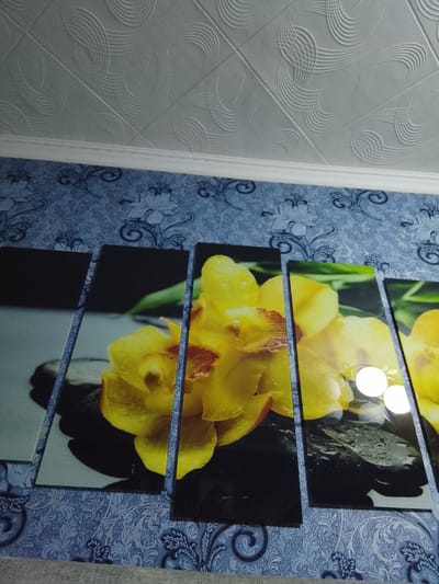 The image shows a wall in a room in Namangan City, Uzbekistan, decorated with a five-panel artwork of yellow orchids and dark stones. The artwork is mounted on a blue patterned wallpaper. The ceiling above the wall has a textured white finish.