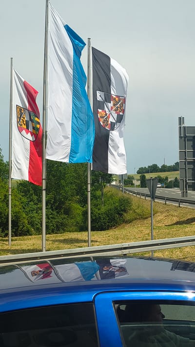 Three flags are displayed on flagpoles along a road in Bayreuth, Germany. The flag on the left is red and white vertically with a shield on the upper hoist side. The middle flag is white with a blue vertical stripe. The flag on the right is black and white vertically with a shield on the upper hoist side. The flags are reflecting on the roof of a blue car in the foreground. In the background, a highway can be seen with trees and vegetation on either side. The sky is overcast.