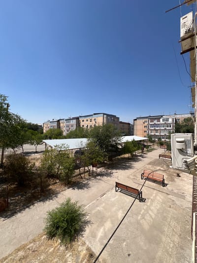 The image depicts a sunny day in a courtyard area in the city of Şamaldı-Say, Kyrgyzstan. In the foreground, a concrete patio area is visible, with two parallel wooden benches with metal frames situated on it. A single green bush is planted in a patch of dry grass to the left of the benches. In the midground, a covered parking or seating area with a light-colored metal roof is present. Beyond this, a row of apartment buildings of varying heights and architectural styles stretches across the horizon under a clear blue sky. These buildings are several stories tall and appear to be made of brick and concrete. Green trees and bushes are scattered throughout the courtyard and surrounding areas, casting shadows on the ground. An outdoor air conditioning unit is mounted on the side of a building on the right edge of the frame. The overall scene conveys a sense of a residential urban environment on a warm, clear day.