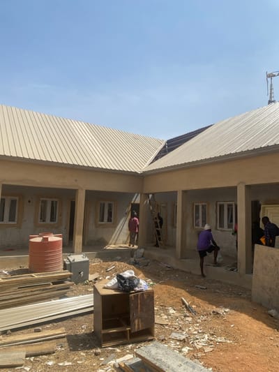 The scene depicts an active construction site within the city of Bisichi, Nigeria, under a clear blue sky. A newly constructed or renovated U-shaped building features a light beige corrugated metal roof, largely complete but with an exposed framework section at the inner corner. The building's exterior walls are plastered in a light beige tone, and multiple white-framed windows are installed in various openings. A covered walkway, supported by pillars, extends around the building's inner perimeter, encompassing a central dirt courtyard.
Five individuals are visible within the scene. In the left wing's doorway, a person in a red long-sleeved shirt and dark trousers stands facing away from the viewer. Near the central corner, a dark-clad individual stands beside a wooden ladder leaning against the wall. On the right, a person wearing a purple top, dark shorts, and a white cap is seen squatting on the edge of the elevated walkway. Further right, two additional individuals are partially obscured: one in a blue and white checkered shirt holding a yellow object, and another behind them.
The courtyard foreground contains various construction materials and debris. On the left, a large orange plastic water storage tank rests near a stack of wooden planks and horizontal metal sheets. A grey electrical enclosure is situated next to the water tank. In the center foreground, a damaged wooden cabinet with open compartments and a black plastic bag on top sits amidst scattered dirt, small concrete fragments, and a metal trowel. The ground throughout the courtyard is uneven, composed primarily of dirt and sand with dispersed debris. A metal tower structure, possibly a street light or antenna, is visible above the roofline on the far right horizon.