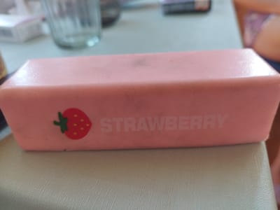 A pink rectangular object, likely an eraser or a small container, with a strawberry graphic and the word "STRAWBERRY" printed on its side, rests on a light-colored textured surface. In the blurred background, a glass and other indistinct objects are visible. The scene appears to be indoors, possibly on a table or desk. The location is reported as Драчкаўскі сельскі Савет, Belarus.
