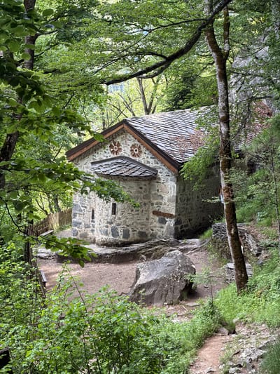 A stone building with a gray tiled roof is nestled in a forest, possibly near a rock face. The building has decorative floral designs on its front façade. A wooden walkway appears to lead to the building. The image may be of the St. George the Victorious Church in Govedartsi, Bulgaria.