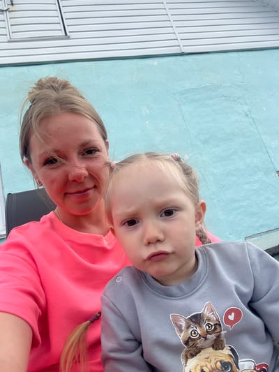 The image features a woman and a young child, likely a mother and daughter, posing for a selfie. The woman is wearing a vibrant pink long-sleeved shirt, and the child is in a grey sweatshirt adorned with a graphic of two cats, one with a red heart bubble. The child's hair is styled in two pigtails. The background consists of a light blue textured wall and a white ribbed structure, possibly part of a building or fence. The lighting suggests it's daytime, with soft, natural light illuminating the subjects. Both individuals appear relaxed and happy, with the child leaning in towards the camera. The image seems to capture a candid, everyday moment outdoors, possibly in a backyard or on a porch, given the rustic nature of the background elements. The overall mood is warm and affectionate, highlighting the bond between the two. There is no visible text in the image.