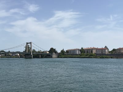 The image shows a wide, calm river flowing through a town. A suspension bridge with a stone support on the left side spans the river, connecting to a distant bank lined with buildings and trees. The buildings are a mix of residential and commercial structures, with several stories and tiled roofs, characteristic of French architecture. The sky above is partly cloudy with a clear blue expanse. The river's surface is rippled, reflecting the overcast sky. The overall scene depicts a tranquil, urban riverside environment in Mercurol-Veaunes, France. There are no people visible in the image.