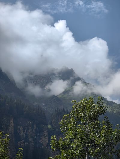 This is a photograph taken in Kothi, India, depicting a mountainous landscape partially obscured by clouds and mist. In the foreground, a leafy green tree is visible, with its branches extending upwards. The middle ground features steep, rugged mountainsides covered in dark green pine trees. Dramatic rock formations are exposed on some of the slopes, adding to the ruggedness of the terrain. Wisps of white clouds and mist drift across the mountains, creating a dynamic and ethereal atmosphere. The background is a clear blue sky with scattered white clouds. The scene conveys a sense of natural beauty and the raw power of the mountains.