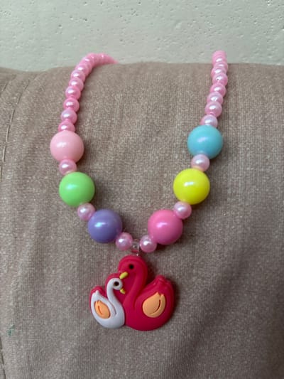 A pink beaded necklace with a swan pendant is displayed on a textured, light brown fabric surface. The necklace features a series of larger, round beads in various pastel colors including pink, green, purple, blue, and yellow, interspersed with smaller pink pearlescent beads. At the bottom of the necklace is a bright pink pendant shaped like two swans, one larger and one smaller, with white detailing on the smaller swan's neck and orange accents. The background is a textured, light-colored wall. The image does not contain any information or clues regarding the city of Imeni Morozova, Russia.