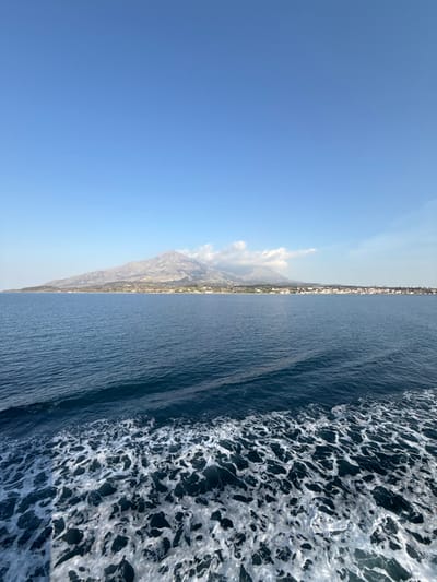 The scene depicts a maritime view of the coastline of Kamariotissa, Greece, under a clear, bright blue sky. A prominent mountain range, characterized by rugged, grayish-brown terrain and sparse vegetation, extends across the horizon, with light cloud formations partially obscuring its peaks. Below the mountains, the town of Kamariotissa is visible as a dense cluster of white and light-colored buildings with reddish roofs, situated along the coastal strip. The foreground consists of deep blue sea, featuring a distinct pattern of white, turbulent foam that indicates the wake of a moving vessel. No individuals or specific vessel components are discernible within the frame. The environment is open and exterior, under daylight conditions.
