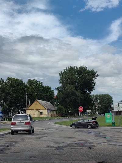 The image shows a street scene in the city of Фаніпальскі сельскі Савет, Belarus. In the foreground, two cars are parked on a damaged asphalt road. The car on the left is a silver minivan with a license plate that reads "E5898 PA-5". The car on the right is a dark-colored sedan with its trunk open. In the background, there is a yellow wooden house with a gray roof, surrounded by lush green trees. A stop sign is visible on a pole near the sedan, along with a street sign that appears to say "ВЯЗАНЬ". Further in the background, a white sign with green lettering is mounted on a pole, possibly indicating a business or service. The sky is partly cloudy with a mix of blue sky and gray clouds. The overall scene depicts a quiet, somewhat rural part of the city on a cloudy day.