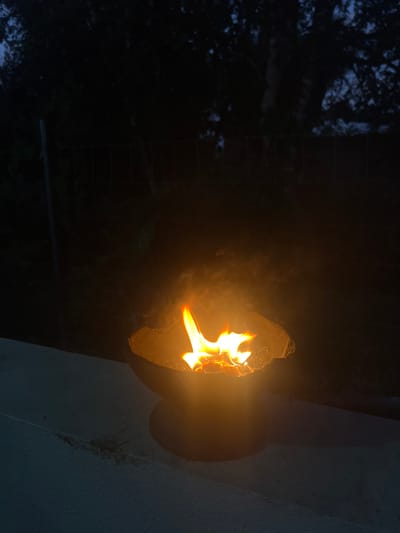 A lit tiki torch is affixed to a white concrete structure in Punta Gorda Town, Belize. The flame emanates from the torch head, casting a warm, flickering glow. To the left, a darkened area suggests the presence of trees or foliage. The background is mostly dark, indicating nighttime.