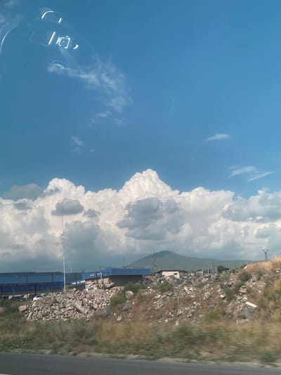 The image depicts a view from a vehicle traveling along a road in the vicinity of Balahovit, Armenia. The scene is dominated by a bright blue sky filled with large, fluffy white cumulus clouds. In the foreground, a dry, grassy embankment littered with numerous rocks and rubble slopes upwards from the road. Beyond the embankment, a blue-roofed building with a long structure extending from it is visible, along with a smaller white building. Power lines and transmission towers are also present in the distance, and a hazy mountain range can be seen in the far background. A reflection of the vehicle's interior, including what appears to be a dashboard and windshield wipers, is visible in the upper left portion of the image, superimposed on the sky. The overall impression is one of a semi-arid, industrial, or semi-developed area under a vast sky.