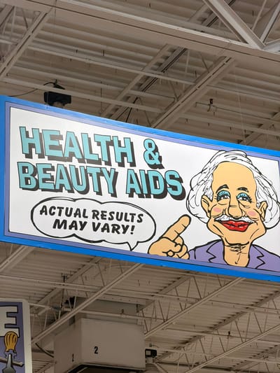 This media file shows a close-up, angled view of a large, brightly colored banner hanging from the ceiling of an indoor venue. The banner features a cartoonish illustration of an elderly woman with bright blue eyes, exaggerated facial features, and white hair. She is wearing a purple top and pointing with her right hand.
Dominating the banner is large, bold text in light blue with a dark blue outline. The text reads "HEALTH & BEAUTY AIDS." To the right of this main text, within a speech bubble graphic, is smaller text in black that says "ACTUAL RESULTS MAY VARY!". The overall tone of the banner is whimsical and attention-grabbing.
The background of the banner is white, and it is framed by a thick blue border. The banner itself is suspended by wires from a complex white metal structure that forms the ceiling of the space. This structure appears to be part of a large building, possibly a warehouse, retail store, or community center, given the industrial nature of the ceiling. Visible in the background, beyond the banner, are more metal beams, corrugated metal siding, and what appear to be HVAC ducts. A white rectangular object with the number "2" printed on it is also visible in the upper right portion of the frame.
The image is well-lit, suggesting an indoor environment without natural light. The orientation of the banner and the perspective of the shot indicate it is a photograph taken from below, looking upwards. The scene is static, with the banner simply hanging as a display or advertisement. There are no people actively engaged in any activity in the frame, only the depicted illustration on the banner. The location is identified as Elizabeth City, United States of America.