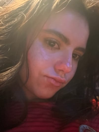 A close-up, tilted portrait features a person, likely female, with long, dark hair. The subject's face is prominent, turned slightly, with dark eyes looking towards the viewer. Her nose is distinctive, and her lips are full with a neutral to slightly contemplative expression. A bright, diffused light source on the left side of the frame causes a significant lens flare effect, washing out part of the image and illuminating the left side of her face and hair with a warm glow. The background is dark and indistinct, suggesting an indoor setting. On the far right, a red garment with subtle striped patterns is visible, along with a small, partially obscured metallic or silver-colored object. The overall impression is one of a casual selfie or portrait taken in low light conditions, possibly while reclining. There is no visible text. The location context provided is College Station, United States of America.