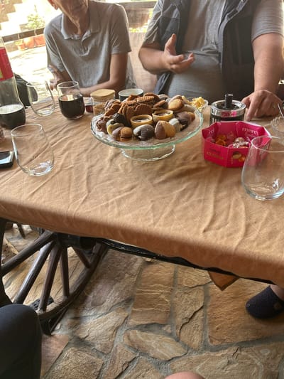 The image depicts a gathering around a table in Bukovlak, Bulgaria. The table is covered with a tan cloth, and it holds a glass bowl filled with assorted cookies and chocolates, a pink box of "Merci Lovelies" chocolates, several wine glasses, a bottle of wine, and an ashtray. Three people are visible partially in the shot. One is wearing a gray shirt and a dark vest, another wears a gray shirt, and a third shows a foot with a slipper. The table appears to be attached to a decorative wooden wheel. The floor is made of stone tiles. The scene suggests a casual, possibly outdoor, setting with people enjoying refreshments.