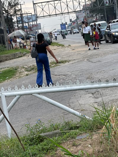 A road scene features a woman in a black top and blue jeans, walking along a metal barrier with triangular spikes. The barrier is located between the road and a patch of overgrown grass. Traffic, including cars and pedestrians, is visible in the background. Billboards line the street. The image is captured at an angle, giving a tilted perspective.