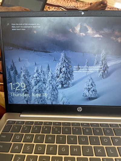 The image displays the lock screen of an HP laptop. The background is a serene winter landscape with snow-covered pine trees and mountains under a cloudy sky. The time "1:29" is prominently displayed in the bottom left corner, followed by "Thursday, June 18". To the left of the time, a search icon precedes the text "From the look of this snowpack, you likely won't be surprised to learn that skiers love it here." On the right side of the screen, another search icon is present, followed by the text "Above the snow-cloaked trees and hills, the clouds part for a peek at a grand peak of the Slovenian Alps." The HP logo is visible on the laptop's bezel below the screen. The keyboard of the laptop is also visible in the foreground. The image is a close-up of the laptop screen and keyboard, taken at an angle. There is no information in the image that suggests the location is Wai A, Nigeria.