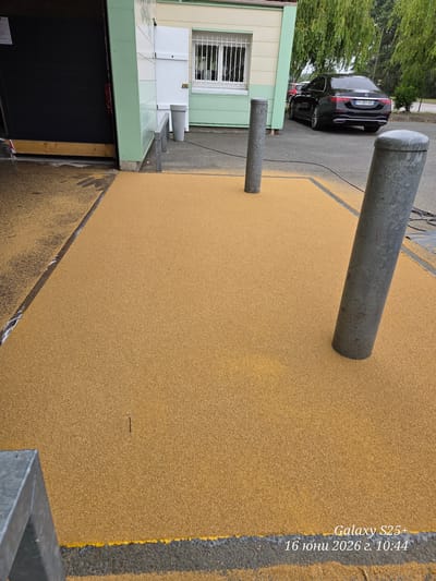 The image depicts an outdoor scene in Les Attaques, France, featuring a newly laid yellow gravel path leading to a building with a green and white facade. The path appears to be wet or recently treated, with scattered yellow granules. Several metal bollards are positioned on and around the path, with two prominent grey cylindrical bollards in the foreground and mid-ground. A black Mercedes-Benz sedan is parked on the asphalt adjacent to the path, identifiable by its license plate "FY-351-GA". The building has windows with metal bars and an open doorway. The overall environment suggests an urban or semi-urban setting, possibly a public facility or entrance area. The image also includes a timestamp in the bottom right corner: "Galaxy S25+ 16 июня 2026 г. 10:44".