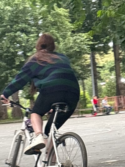 The image shows a person riding a bicycle in an outdoor setting. The person is wearing a striped sweater and dark pants. The bicycle is white and has a visible front wheel and handlebars. The background consists of lush green trees and foliage, suggesting a park or natural area. In the distance, there are some structures that appear to be benches or picnic tables, with other people visible sitting on them. The lighting suggests it is daytime, and the presence of abundant greenery indicates a warm weather season. The image is taken from a low angle, looking up at the cyclist.