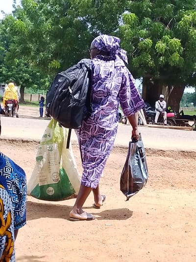 The image shows a woman walking away from the camera, carrying a large black backpack on her shoulders and a black shopping bag in her right hand. She is wearing a purple and white patterned outfit and a matching headscarf. The ground is sandy and reddish-brown, suggesting a dry, possibly rural environment. Behind her, a large, light-colored bag with green accents is visible. In the background, there are trees with green foliage, a road, and a few other people. One person is on a motorcycle, wearing a yellow garment. Another person, a child in blue, stands near the road. Further in the background, a man in white sits on a motorcycle under a tree. The scene appears to be outdoors during daylight, likely daytime, with overcast conditions suggested by the lighting. There are no obvious indicators of the specific time of day beyond it being daylight. The location is stated as Oke Iho, Nigeria. Some text is visible on the light-colored bag: "25Kg". The black shopping bag has some white text that is partially visible. The overall impression is of a person on the move, carrying belongings in a public outdoor space.