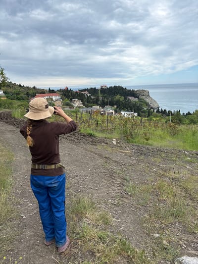A woman with a long blonde braid, wearing a tan hat, brown jacket, blue pants, and dark shoes, is standing on a dirt path, holding a camera in her hands. She is positioned to the left of the frame, facing towards a coastal scene.
In the background, there is a view of a town or city nestled along the coast of the Black Sea. There are buildings, trees, and what appears to be a rocky promontory extending into the water. The sky is filled with gray and white clouds.
The environment is dominated by the natural landscape, with vegetation and the dirt path visible in the foreground, while the city of Ut'os, Ukraine, is present on the coast in the background. The woman seems to be taking a photograph of the scenery.