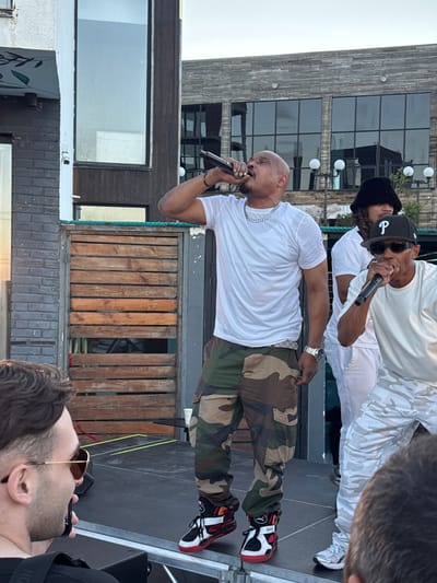 In this outdoor scene set in Mukhatskaro, Georgia, two men are the central subjects. One man, with a shaved head and beard, stands center-left, wearing a white t-shirt, a silver chain, and camouflage pants. He is holding a microphone to his mouth, appearing to be singing or rapping. On his right wrist, he wears a silver watch. His footwear consists of red, black, and white athletic shoes.
Behind him and slightly to the left, another man, wearing a black beanie and sunglasses, is also holding a microphone. He is dressed in a white t-shirt and white camouflage pants. Both men are performing on a raised platform with a dark-colored surface. In the background, a building with large windows and a stone facade is visible. Further back, a structure made of vertical wooden planks can be seen. The lighting suggests it is daytime, possibly late afternoon or early evening. The overall mood appears energetic, consistent with a live musical performance.