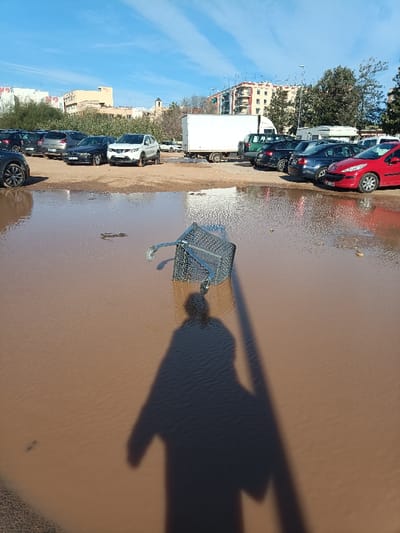 The image displays an outdoor scene in what appears to be an unpaved parking area in Burjassot, Spain, under a clear blue sky with scattered clouds. The foreground is dominated by a large, irregularly shaped puddle of reddish-brown muddy water. Submerged partially in this water, slightly right of center, is a metal shopping cart lying on its side. The elongated shadow of a person, likely the photographer, is cast across the muddy water, extending from the bottom center towards the middle right of the frame.

Behind the puddle, numerous vehicles are parked on a drier, unpaved surface. These include several passenger cars of various colors (dark grey SUV, dark blue sedan, silver hatchback, red hatchback), a white box truck with a dark undercarriage, and a green van. In the distant background, several multi-story residential buildings with terracotta roofs are visible. A prominent older building with a bell tower or spire can be seen on the far left horizon, suggesting a civic or religious structure. Scattered trees with sparse foliage are also present between the parked vehicles and the buildings. The scene is illuminated by direct sunlight, indicated by distinct shadows.