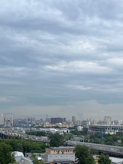This is a vertical, high-angle, full shot of a city under a cloudy sky. The sky is filled with a thick layer of gray clouds, obscuring the sun and creating a diffused, dim light across the entire scene. In the foreground, the roof of a building with a white and gray facade is visible, with some surrounding greenery and other industrial-looking buildings with white roofs and gray walls.
The midground is dominated by a multi-level highway system with moderate traffic, including cars and trucks. The highway is surrounded by lush green trees. Beyond the highway, a sprawling urban landscape unfolds, featuring a variety of buildings ranging from low-rise structures to tall, modern skyscrapers. A large stadium with white, arching supports is prominently visible in the right midground. Industrial buildings and smokestacks can be seen throughout the city. The overall impression is that of a densely populated, active city.
The image appears to be a cityscape of Tarasovka, Russia, judging by the distinct architecture and the presence of what looks like the Arena CSKA stadium.