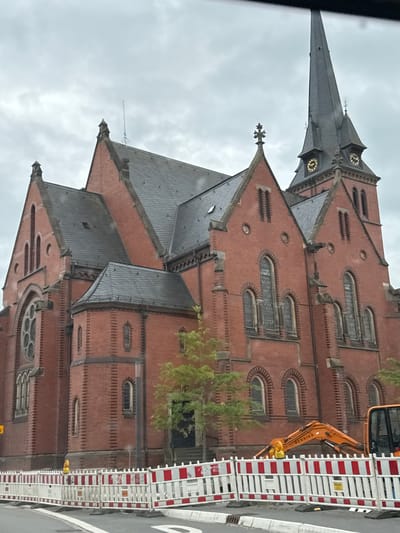 An overcast day frames a large, red brick church with a tall, dark grey, slate-covered spire. The church has multiple gabled roofs and arched windows. The spire features two clock faces with gold numerals. In front of the church, construction barriers with red and white stripes cordon off an area. An orange excavator is visible behind the barriers, and a yellow construction cone is also present. Trees with green foliage are to the right of the church. The scene is located in Gronau, Germany.