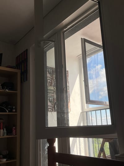 The image depicts an interior view of a room with a window leading to a balcony. In the foreground, a bookshelf is visible on the left, containing various items. A painting with a colorful abstract pattern hangs on the wall above the bookshelf. The main focus of the image is the window, which is partially open, revealing a view of a balcony with a railing. Beyond the balcony, a bright blue sky with white clouds is visible. In the lower part of the image, through another window, a wooden ladder and greenery can be seen, suggesting an outdoor environment. The overall lighting suggests it is daytime, with sunlight casting shadows in the room. The scene appears to be a residential interior.