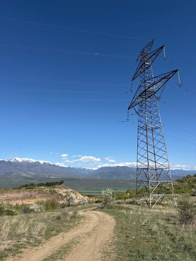 The image depicts a tall, steel lattice transmission tower standing in a grassy field, adjacent to a dirt path. The tower extends vertically into a clear, blue sky with power lines running parallel to each other, spanning the scene. In the background are mountain ranges with patches of snow visible on their peaks. Patches of green vegetation and some white flowering trees dot the landscape at the foot of the mountains. The location, as requested, is Tsarvishte, Bulgaria. No people or interactions are visible in the image.