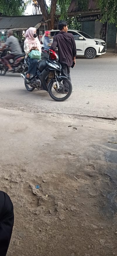 In Gampongteungoh, Indonesia, a young woman wearing a light pink hijab and a black face mask is seated on a black motorcycle. She is holding a light green plastic bag in her lap and has her right hand on her phone. Beside her, a young man in a dark long-sleeved shirt and dark pants stands, holding his phone in his right hand. The motorcycle has red accents on the front and silver spokes on the wheels. In the background, a white car is parked, and a building with a sign that appears to read "Pangkas Su" is visible. Trees with lush green foliage line the street, and another person on a motorcycle is blurred in motion to the left. The foreground shows a rough, textured concrete surface with some debris and tire tracks. The scene appears to be a roadside setting, possibly near a shop or business.