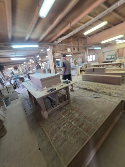This is a wide-angle, low-resolution photograph of the interior of a workshop, likely in Kaleytsa, Bulgaria. The scene is a long, narrow space with a predominantly wooden interior, illuminated by fluorescent strip lights running along the ceiling. The workshop appears to be under construction or renovation, as evidenced by exposed wooden beams and unfinished walls.
The floor is a mix of brick and concrete, with a significant amount of dust and debris, suggesting active work is in progress. In the foreground and midground, there are various wooden furniture pieces, some partially assembled or in components. Tools, including what appears to be a power tool, are scattered on a workbench. A person, wearing a grey shirt and dark shorts, is partially visible, possibly working on a piece of furniture. The person's face is not clearly visible.
Towards the back of the workshop, more unfinished wooden structures and materials are visible. The lighting suggests it could be daytime, but the interior setting makes it difficult to determine the exact time of day. There are no clear indications of the weather outside. The overall atmosphere is one of a busy, functional workshop environment. No visible text is present in the image.