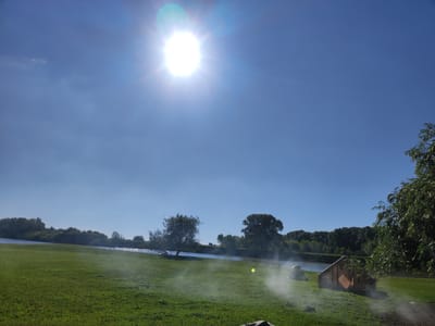 The image depicts a sunny day at a grassy park beside a body of water, likely the river in Архангель ауылдық округі, Kazakhstan. The sun is high in the bright blue sky, causing a lens flare effect. In the foreground, a green lawn is visible, with a small wooden structure resembling a playhouse or a shed on the right side. To the left of this structure, there appears to be a tent, and a white misty substance, possibly smoke or fog, is present on the grass between the structures and the water. Several trees are scattered across the landscape, with a dense line of trees forming the background along the opposite bank of the water. The water itself reflects the sunlight. No people are visible in the scene. The overall impression is one of a natural outdoor setting on a clear day.