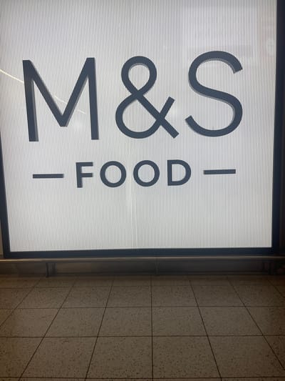 The image displays a brightly illuminated retail sign, positioned vertically, with its left side featuring the text "M&S" and its right side reading "- FOOD". The text is rendered in dark grey against a white background that has subtle horizontal line patterns, suggesting a ribbed or textured surface or an effect of the backlighting. The sign is enclosed within a dark frame.
To the right of the sign, a light beige or off-white tiled wall is visible, consisting of rectangular tiles with darker grout lines. The tiles appear to have a slightly speckled texture. No people or specific activities are depicted in the image. The lighting on the sign is prominent, indicating that it is either actively lit or in a well-lit indoor environment.
The text "M&S - FOOD" clearly identifies the sign as belonging to a Marks & Spencer Food store, a well-known British retailer. Given the provided context, the image was captured in Horley, United Kingdom. The scene suggests an interior space, likely a shopping area or a walkway adjacent to a store.