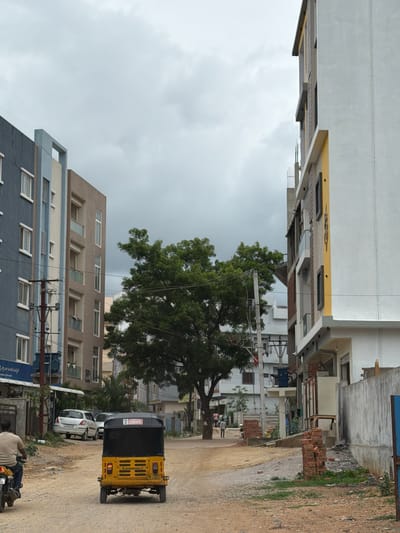 An overcast, daytime shot captures a street scene in Shamshabad, India, featuring a mix of residential and commercial buildings, vehicular traffic, and pedestrian activity. On the left, a blue and gray multi-story building stands next to a beige and white building. A power pole with multiple wires is situated in front of these structures. To the right, a prominent white and yellow building, also multi-story, is under construction or recently completed, with exposed brickwork and scaffolding visible in some areas. A large, leafy green tree is centered in the mid-ground, partially obscuring a smaller white building behind it. A yellow auto-rickshaw is parked on the dirt road in the foreground, facing away from the camera. To the left of the auto-rickshaw, a person on a motorcycle is moving away from the viewer. In the background, several cars are parked near a building, and a few pedestrians are walking on the right side of the street. The road surface is primarily dirt with some gravel and scattered bricks. The sky is cloudy and grey.