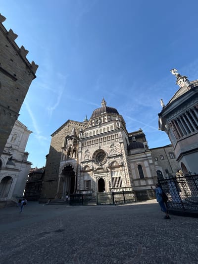 The image depicts the Piazza del Duomo in Bergamo, Italy, bathed in bright sunlight under a clear blue sky with a few wispy clouds. The scene is dominated by the impressive facade of the Basilica di Santa Maria Maggiore, characterized by its ornate decorations, large rose window, and a central dome. To the left, the side of another building, possibly a church or bell tower, with a crenellated top, is visible. On the right, a portion of a structure with classical columns and statues can be seen.

The ground of the piazza is paved with cobblestones. Several people are present in the scene. A person wearing a blue shirt and shorts is standing near a black wrought-iron fence, holding up a phone as if taking a picture. Further in the background, near the entrance of the basilica, a few individuals are seen walking or standing. On the left side of the image, two people are walking away from the camera towards a white building with arched entrances. The overall atmosphere is that of a sunny day in a historic Italian city, with tourists exploring the architectural landmarks.