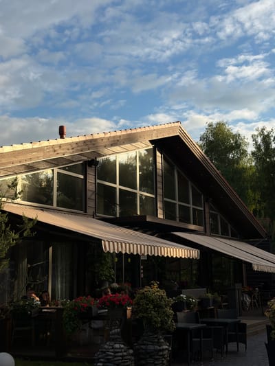 A low-angle shot depicts the exterior of a wooden, A-frame structure with large windows, likely a restaurant or cafe, in Polivanovo, Russia. The building features an awning with a striped pattern. Outdoor seating with dark chairs and tables is visible to the right, decorated with flowers and plants. The reflection of trees and sky can be seen in the windows. The sky is blue with scattered clouds.