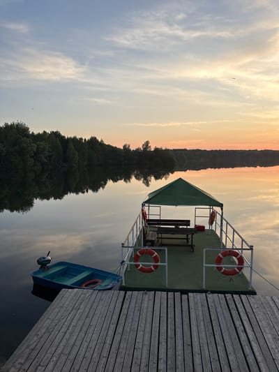 The image depicts a serene lake scene at dusk, likely near Kamysh, Russia. The sky is painted with soft hues of orange, yellow, and blue, with wispy clouds reflecting on the still water. A wooden pier extends into the lake, with a small blue motorboat tied to it. Further out, a green-roofed floating platform features a picnic table with benches and several life preservers. The opposite shore is lined with a dense forest, its dark silhouette contrasting with the colorful sky and its reflection mirrored in the water. There are no people visible in the frame.