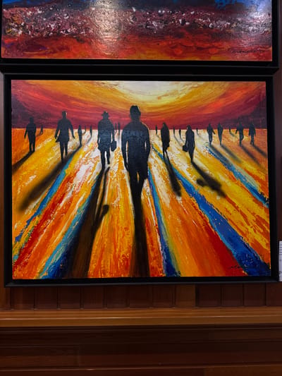 The image displays two framed paintings hanging on a dark wooden wall. The top painting is partially visible and appears to be an abstract depiction of a turbulent, reddish-purple sky. The larger, bottom painting features a group of silhouetted figures walking away from the viewer towards a vibrant, sunset-like sky that transitions from fiery red to yellow. The ground is rendered with thick, textured strokes of orange, yellow, blue, and white, creating a sense of depth and movement. Long, dark shadows stretch out from each figure, contributing to the dramatic atmosphere. The overall style is expressive and colorful, with a focus on conveying emotion through bold brushwork and contrasting hues. A small white label is attached to the lower right corner of the bottom painting's frame, though its text is largely illegible. The paintings are displayed in a well-lit indoor environment, likely an art gallery or exhibition space. There is no indication in the image that this scene is located in Torrejón de Ardoz, Spain.