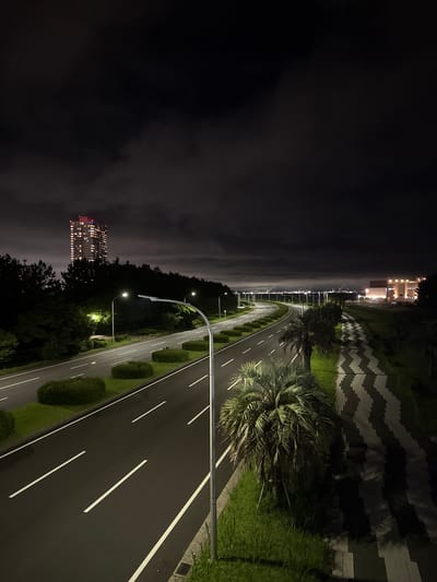 The image depicts a nighttime scene on a multi-lane road, likely a highway or major thoroughfare, within the city of Chiba, Japan. The road is empty of vehicles and illuminated by streetlights and the lights of distant buildings. On the left side of the road, there are manicured bushes and trees, and in the distance, a tall residential building with many lit windows stands out against the dark sky and surrounding trees.

To the right of the road, a wide pedestrian walkway with a distinctive black and white zigzag pattern is visible. This walkway is lined with palm trees and grass. Further in the distance, along the coast, a cluster of buildings with illuminated windows and what appear to be industrial structures emitting smoke or steam can be seen. The sky is dark with some clouds, creating a moody atmosphere. The road surface appears wet, suggesting recent rain. The overall scene conveys a sense of urban development adjacent to a coastal or park-like area at night.