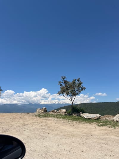 The image shows a landscape with a dirt road on the right side and a clear blue sky on the left. A lone tree stands between the road and a mountain range partially obscured by large white clouds. Behind the mountain, a green hillside is visible. The bottom right corner shows a side mirror. The image appears to be taken in a natural setting. It is not possible to determine the specific location in Leshten, Bulgaria.