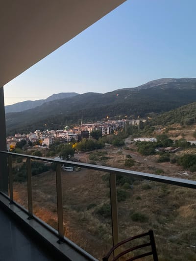The image shows a view from a balcony overlooking a town nestled at the base of a mountainous region. The town is characterized by a mix of residential buildings, including apartment blocks and smaller houses, with some illuminated windows indicating it is dusk or early evening. A few vehicles, including a white van and a yellow car, are visible on a dirt road. The surrounding landscape is hilly and covered in dry vegetation and scattered trees. In the distance, the mountains are covered in dark green foliage. The sky is a clear, pale blue, with a hint of orange near the horizon, suggesting either sunrise or sunset. Two hot air balloons are visible in the sky above the mountains, one with red and white markings. The balcony has a modern metal and glass railing.