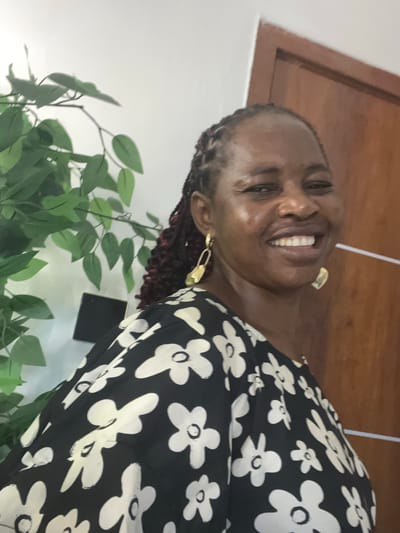 A middle-aged, dark-skinned woman is smiling in front of a light gray wall with a wooden doorframe. She is wearing a black floral dress with white flowers, and gold earrings. The woman has braided hair with red highlights. A green leafy plant is visible in the lower left corner of the image. The location is presumed to be in a building in the city of Mopo-Akinlade, Nigeria.