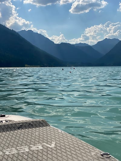 The image depicts a serene scene on a body of water, likely a lake, with mountains in the background. The perspective is from a paddleboard, with its textured surface and a metal handle visible in the foreground on the right. The water is a calm, turquoise color with gentle ripples reflecting the sunlight. Several buoys, appearing as red spheres, are scattered across the water, suggesting a designated swimming or boating area.

In the distance, imposing mountains rise, their slopes covered in dark green forest. The sky above is a vibrant blue, dotted with fluffy white clouds, some of which are casting shadows on the mountains. The sunlight suggests it is daytime, likely late morning or afternoon, with clear weather.

The overall mood is peaceful and recreational. The presence of the buoys and the paddleboard implies that people are engaging in water activities such as swimming, paddleboarding, or perhaps kayaking. No people are explicitly visible in the frame, but the context suggests a leisure outing. There is no visible text in the image. The location is identified as Lärchenwiese, Austria, which aligns with the mountainous and lake scenery.