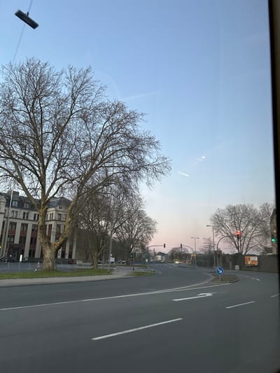 The image depicts an urban street scene at dusk in Hagen, Germany, viewed from within a moving vehicle through a window, indicated by reflections of a wiper blade and light sources. The specific location is the intersection of Frankfurter Straße and Volmestraße/Buscheystraße, with the Kreisverwaltung Hagen (Hagen District Administration) building prominently on the left.

The sky exhibits a clear gradient from light blue overhead to a soft pink-orange tint near the horizon. Multiple contrails are visible in the upper sky. The foreground shows dark asphalt pavement with white lane markings, including a solid line on the left and a dashed line on the right, defining travel lanes. A white arrow painted on the road surface indicates a rightward curve.

On the left, a large, multi-story building with a light-colored facade, numerous windows, and a dark roof, identified as the Kreisverwaltung Hagen, stands behind a green lawn and several mature, bare deciduous trees. Parked vehicles are partially visible near the building's entrance. The wide, multi-lane road curves to the right, while another road branches off to the left. Traffic lights are present; one on the right displays a red signal, and another further left appears green. Metal streetlights line the roadway. A blue circular sign with a white arrow pointing down and to the right, indicating a mandatory direction, is situated on a small traffic island in the mid-right. In the far background on the right, a large billboard displaying an advertisement with orange and brown tones is partially visible, mounted on a low wall. No individuals or active vehicle interactions are discernible in the scene.