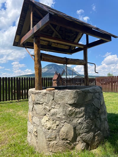 An outdoor shot shows a stone well with a wooden roof and crank mechanism, situated in a grassy area. A wooden fence encloses the property, and in the background, a mountain is visible under a bright blue sky with scattered clouds. A small decorative windmill is placed on the well's stone base.