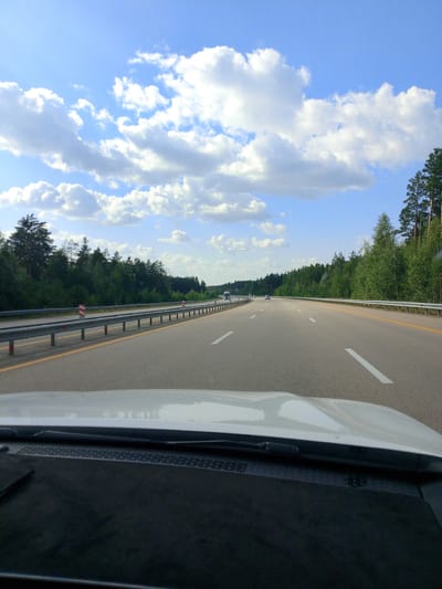 The image shows a view from inside a car driving on a highway. The highway is paved and has clear lane markings. There are guardrails along the sides of the road, with striped posts. On either side of the highway, there are dense forests of green trees. The sky is blue with scattered white clouds. There are several cars visible on the highway in the distance, suggesting traffic. The foreground of the image shows the car's hood and dashboard. The image does not provide enough information to determine the specific location within Алтынды ауылдық округі, Kazakhstan.