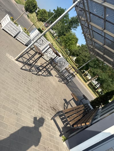 The image depicts an outdoor seating area at "Cool Cafe" in Ivatsevichy, Belarus. Several wooden planter boxes with round topiaries and small yellow flowers are lined up on a paved walkway. Adjacent to the planters are high-top tables with metal frames and wooden seats, also bearing the "Cool Cafe" logo. A wooden bench with a metal frame is positioned in the foreground, casting a shadow on the pavement. Beyond the seating area, a street is visible, lined with trees and grassy areas. A structure with a corrugated metal roof and exposed framing, possibly part of the cafe's outdoor dining space, extends into the frame. The sky is clear and blue, indicating a sunny day. The image is captured from a slightly elevated angle, looking down and across the scene.