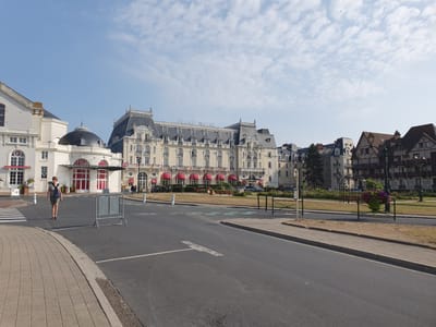 A street view of the Grand Hotel in Cabourg, France, under a partly cloudy sky. A woman wearing a patterned shirt and shorts, with a blue backpack, walks across a crosswalk on the left side of the image. In the background, the Grand Hotel, a large, ornate building with a grey mansard roof and cream-colored facade, is visible. Red awnings cover many of the windows. To the right of the hotel are several half-timbered buildings. A road runs through the foreground, with a sidewalk and a grassy area to the right. Several streetlights and decorative metal fences are also present. Cars are parked in front of the hotel.