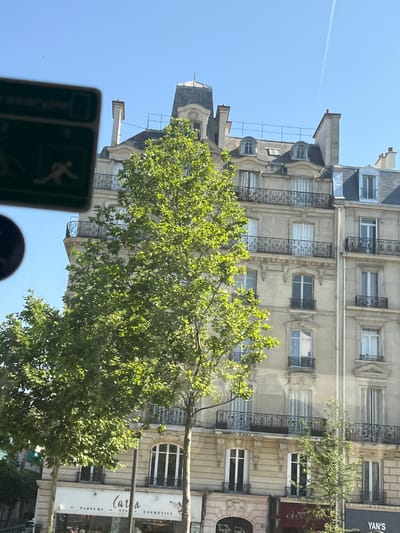 The image displays a building facade in Neuilly-sur-Seine, France, bathed in bright daylight. The building exhibits classic Haussmannian architecture with multiple stories, balconies with ornate railings, and arched windows. Lush green trees with dense foliage partially obscure the lower floors of the building, with their branches reaching towards the clear blue sky. A sign is visible at the bottom of the image, partially obscured by a tree, with text that appears to read "CARLA," "PARFUMS," "SPA," and "COSMETICS." The upper portion of the image captures a faint airplane contrail against the sky. The scene is static and lacks any discernible human activity or interaction.